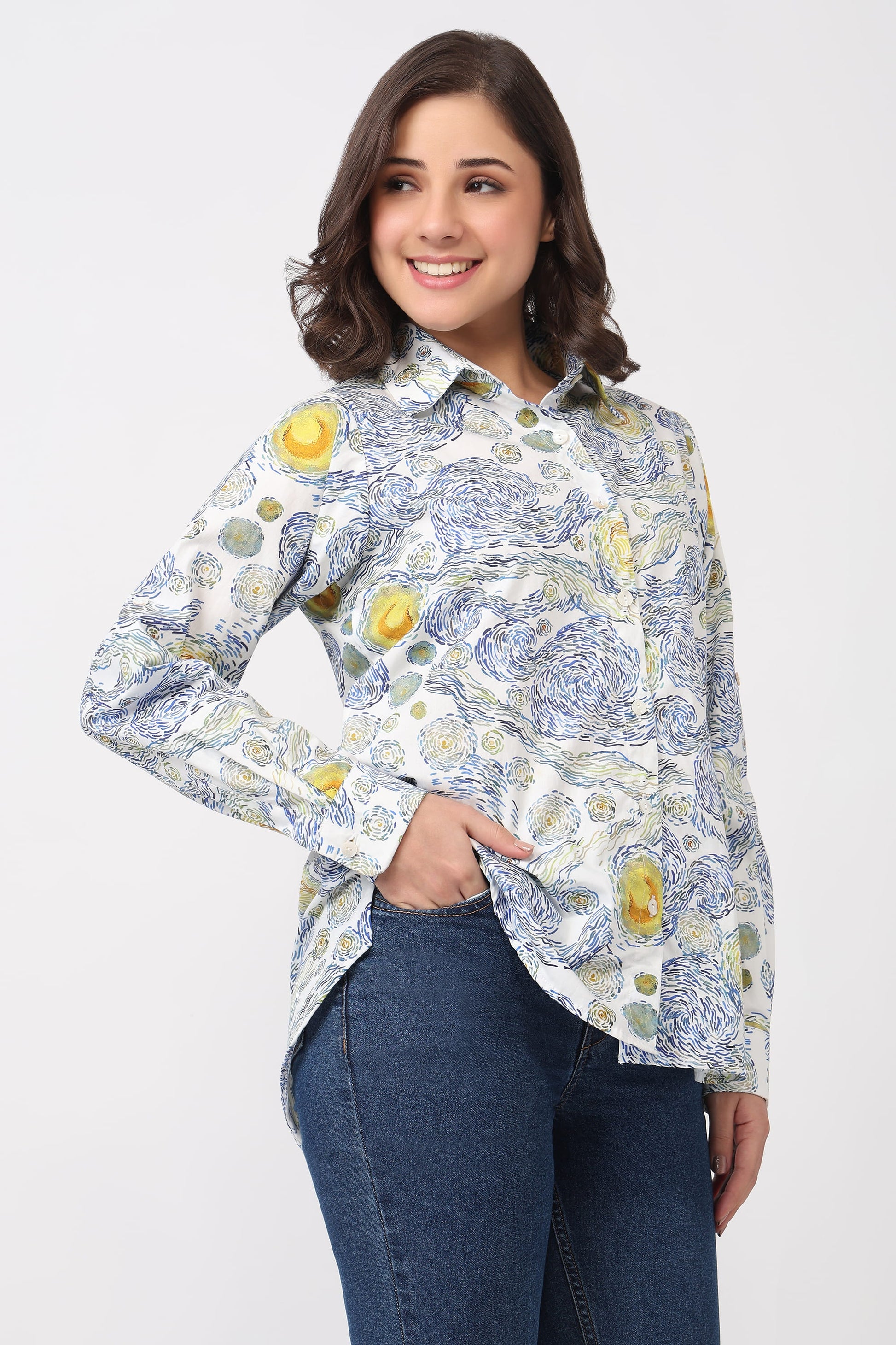 A woman modeling a long-sleeve button-up shirt with a blue and yellow floral print on a white background, paired with dark blue jeans, against a light gray studio background.