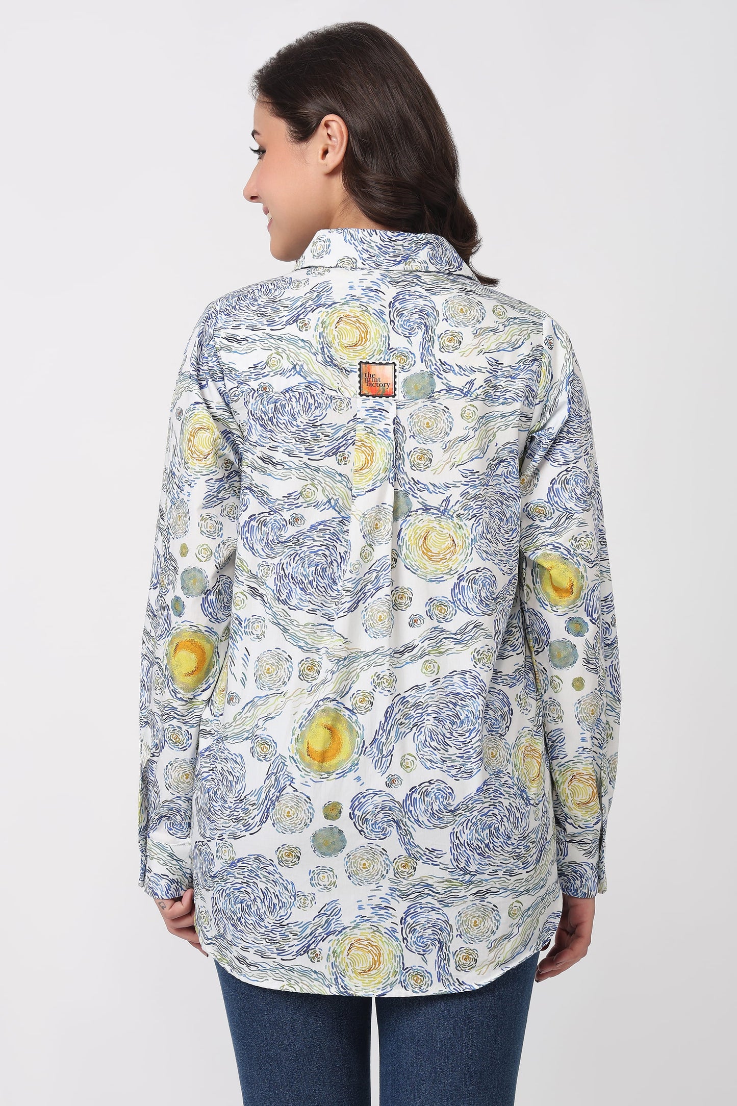 A woman shown from the back wearing a long-sleeve button-up shirt with a blue and yellow floral toile-style print on a white background, paired with blue jeans against a light gray studio background.