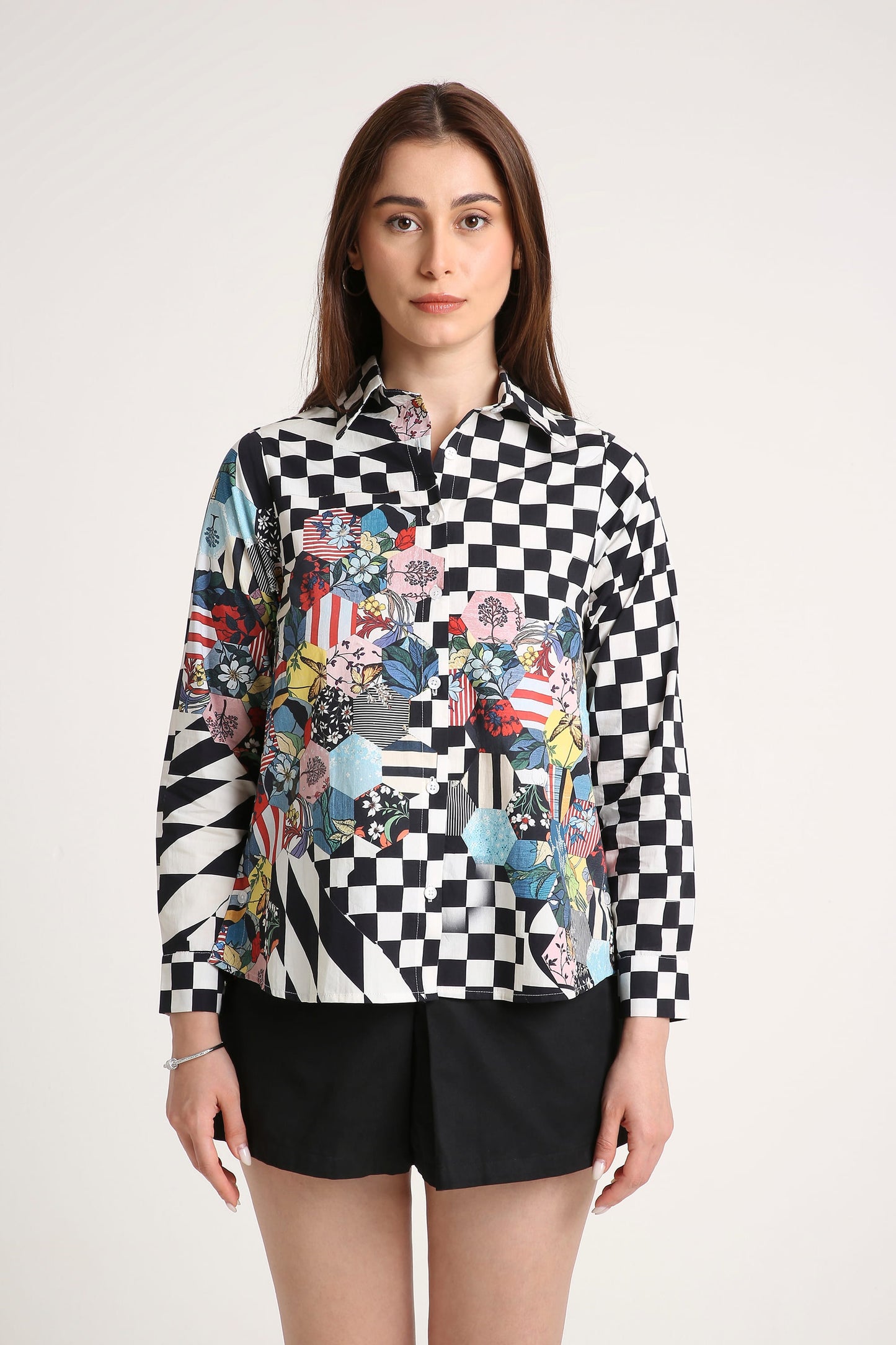 A woman modeling a long-sleeve button-up shirt with a black and white checkerboard pattern on the upper body and sleeves, transitioning into a multicolor patchwork print with floral, geometric, and flag motifs across the lower front. She is wearing black shorts against a plain light gray studio background.