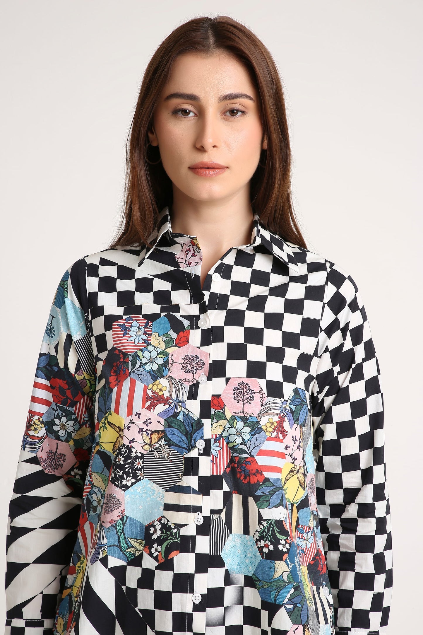 A woman wearing a long-sleeve button-up shirt with a black and white checkerboard pattern on the upper body and sleeves, transitioning into a colorful patchwork print with floral, geometric, and patriotic motifs on the lower front, against a plain light gray background.