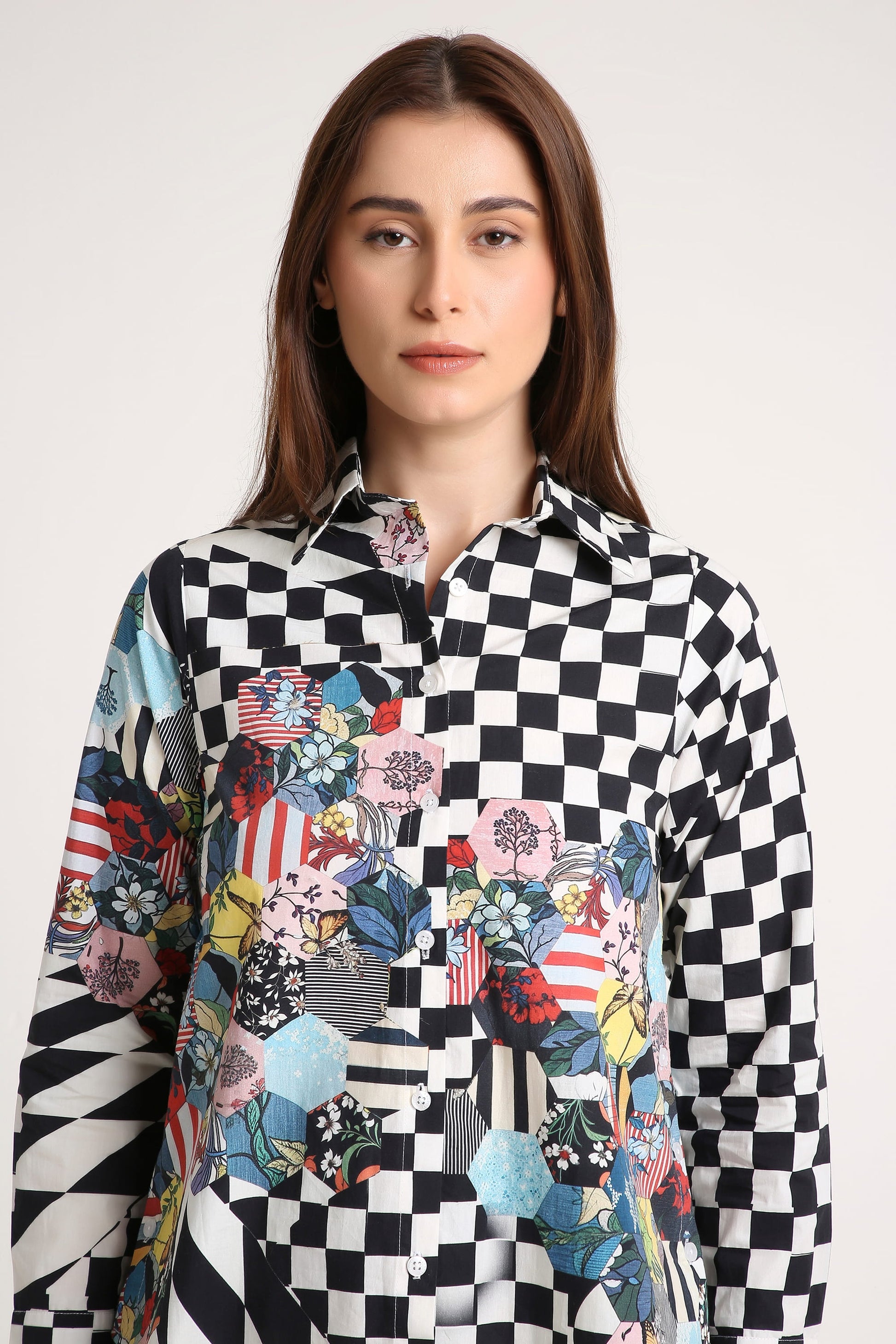 A woman wearing a long-sleeve button-up shirt with a black and white checkerboard pattern on the upper body and sleeves, transitioning into a colorful patchwork print with floral, geometric, and patriotic motifs on the lower front, against a plain light gray background.