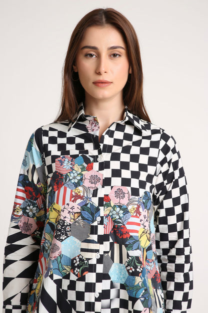 A woman wearing a long-sleeve button-up shirt with a black and white checkerboard pattern on the upper body and sleeves, transitioning into a colorful patchwork print with floral, geometric, and patriotic motifs on the lower front, against a plain light gray background.