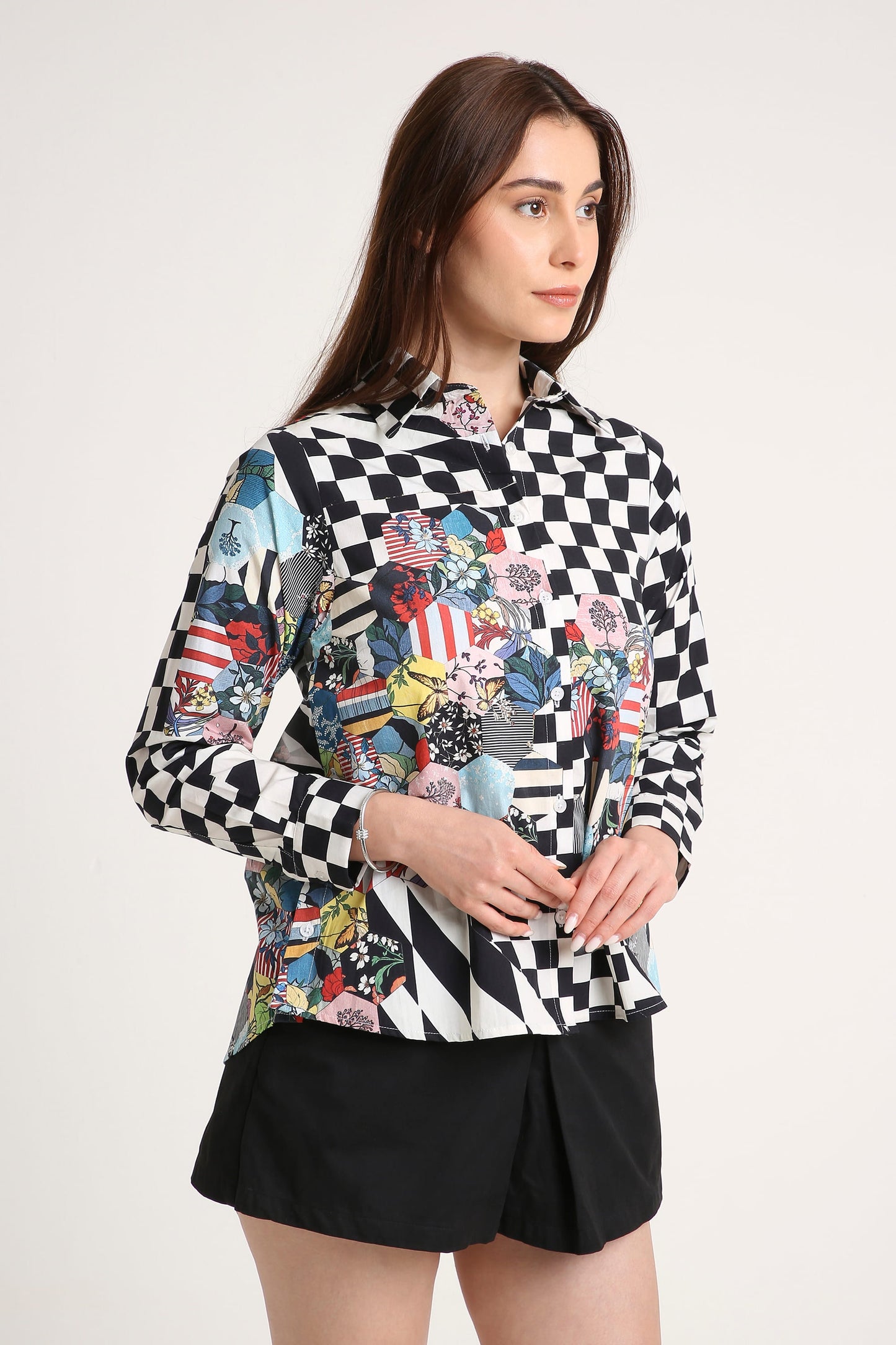 A woman modeling a long-sleeve button-up shirt with a black and white checkerboard pattern on the sleeves and a multicolor abstract collage print on the body, paired with black shorts against a light gray studio background.
