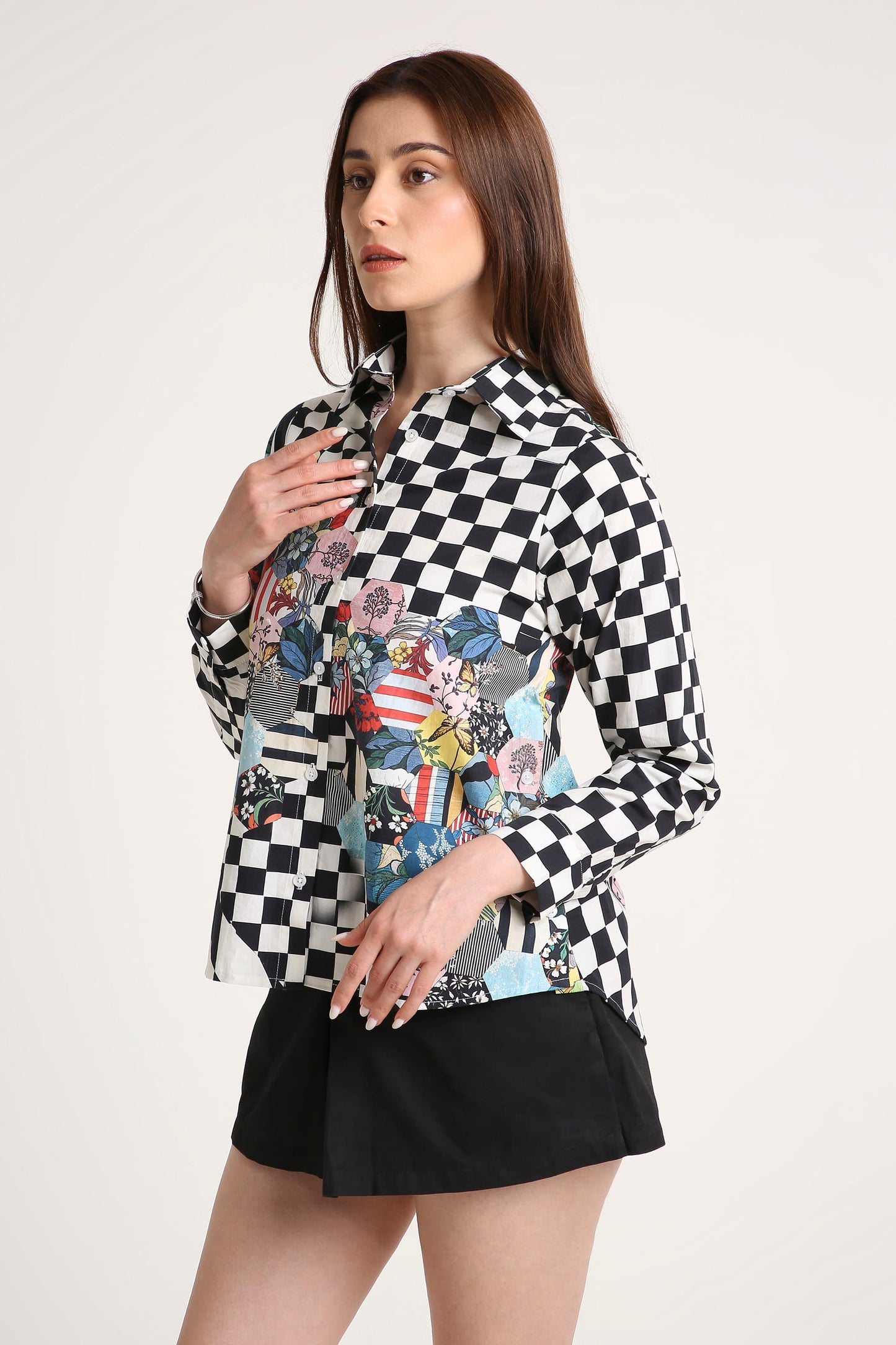 A woman modeling a long-sleeve button-up shirt with a black and white checkerboard pattern on the upper body and a colorful floral patchwork print on the lower front, paired with black shorts against a plain light background.