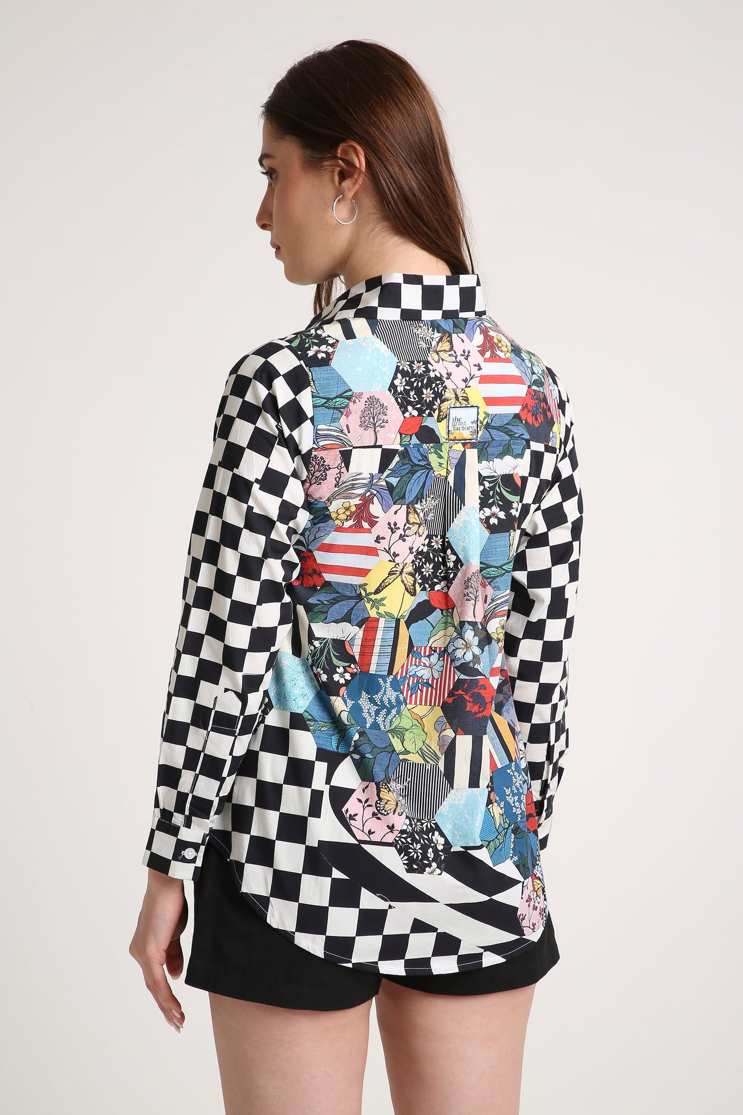 A woman shown from the back wearing a long-sleeve button-up shirt with a black and white checkerboard pattern on the sleeves and a large multicolor abstract collage print across the back, paired with black shorts against a light gray studio background.