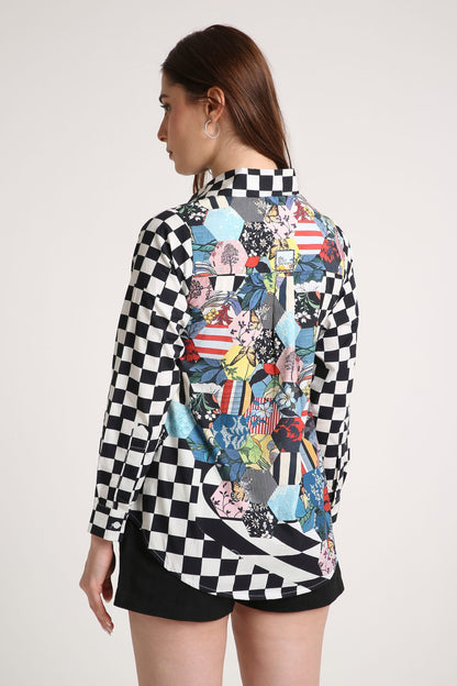 A woman shown from the back wearing a long-sleeve button-up shirt with a black and white checkerboard pattern on the sleeves and a large multicolor abstract collage print across the back, paired with black shorts against a light gray studio background.