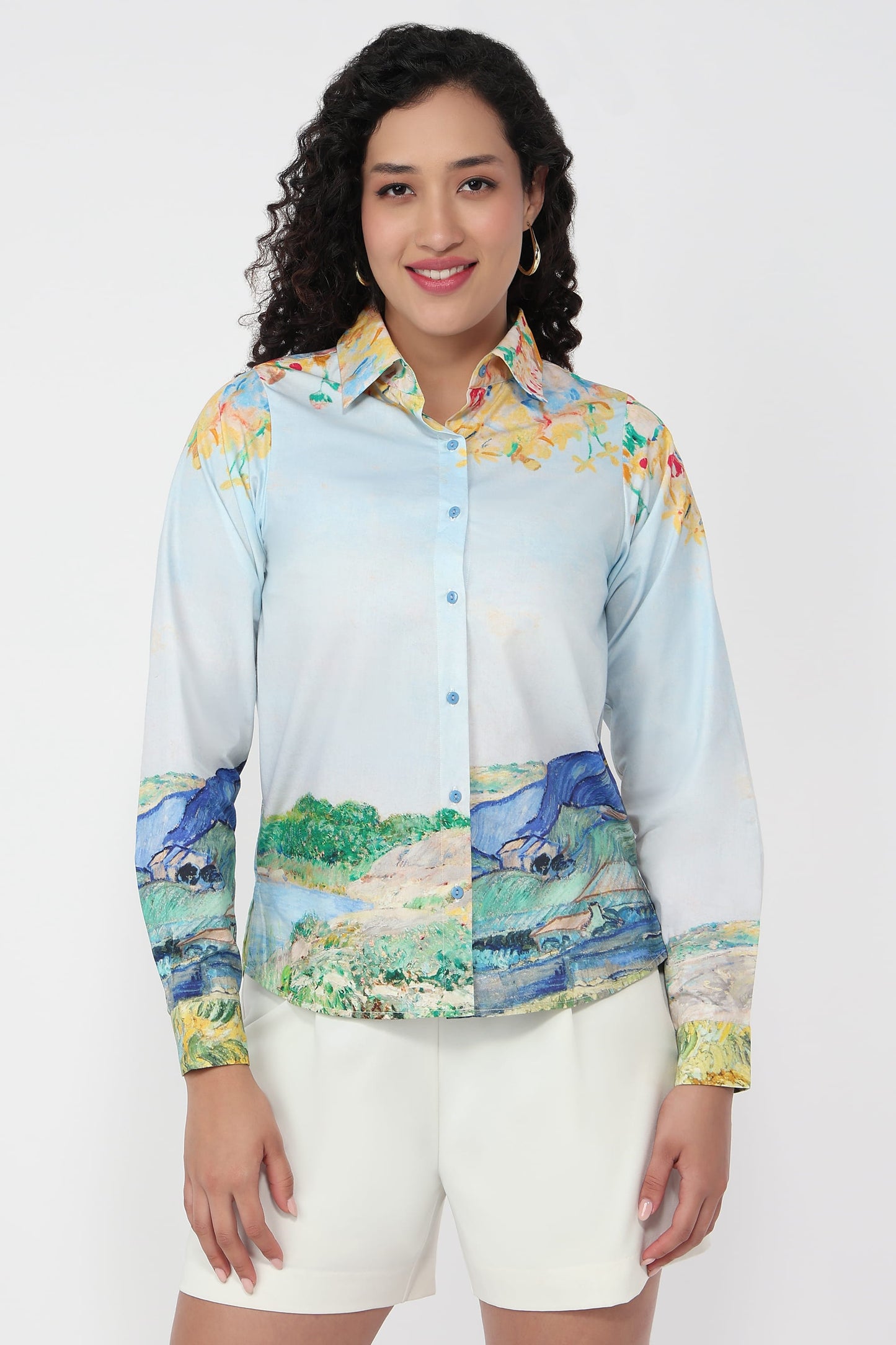 A woman modeling a long-sleeve button-up shirt with a light blue base and a multicolor landscape print on the collar, cuffs, and hem, paired with white shorts against a plain white background.