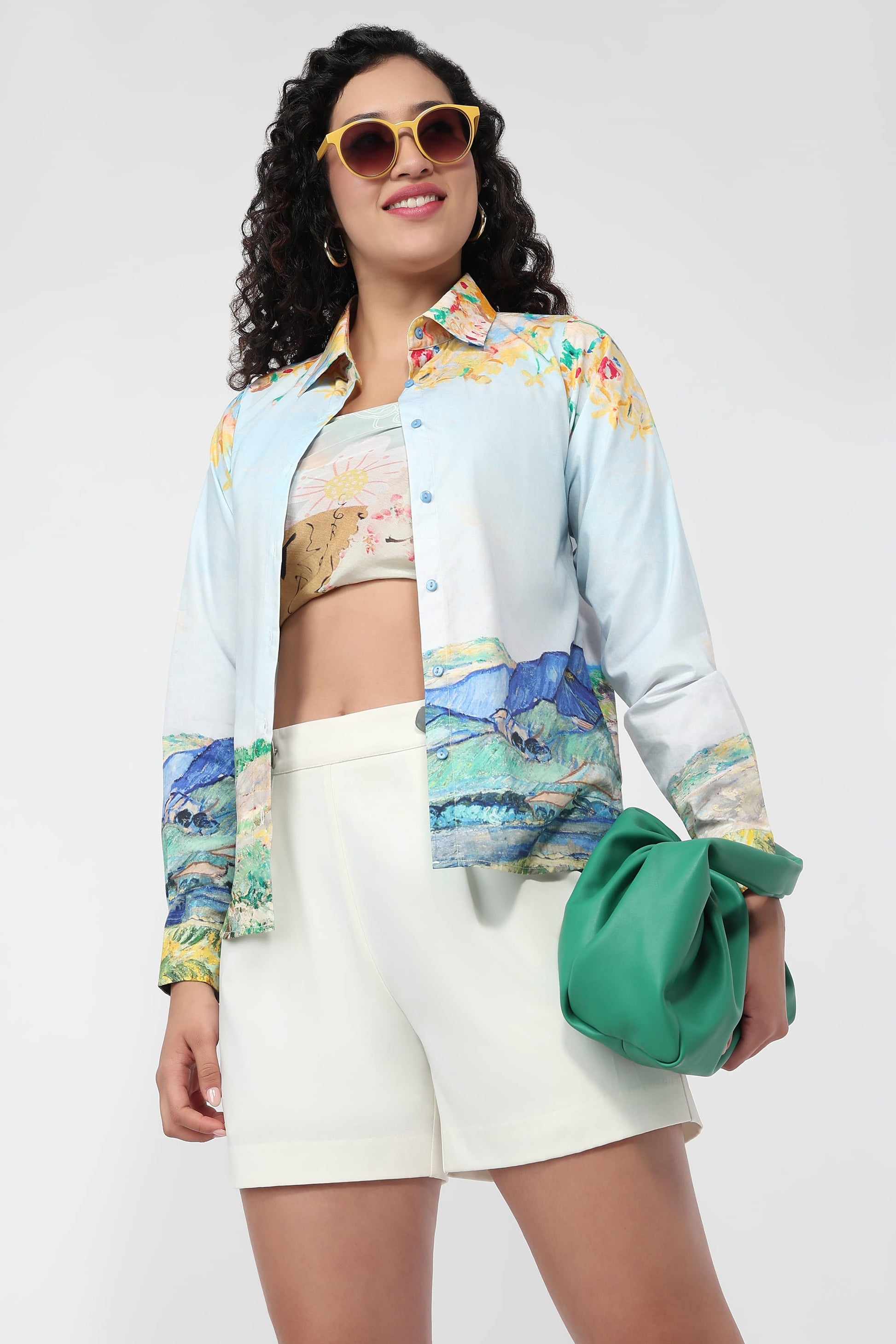 A woman models a light blue button-up shirt with a colorful scenic print on the sleeves and hem, worn open over a matching printed crop top, paired with white shorts and a green clutch bag against a light gray background.