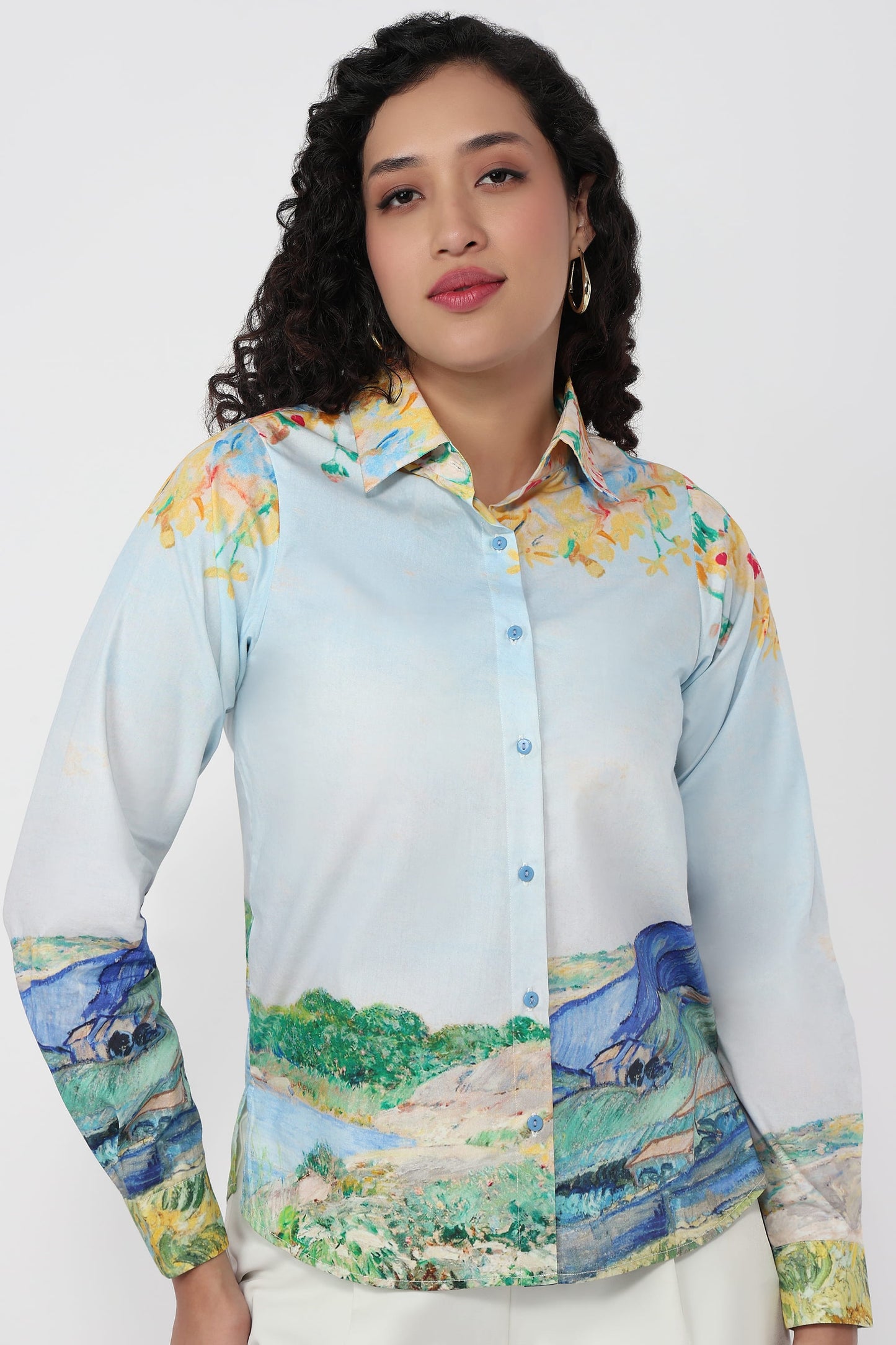 A woman wearing a long-sleeve button-up shirt with a light blue base and a multicolor landscape print featuring horses and green fields, paired with white pants against a plain light gray background.