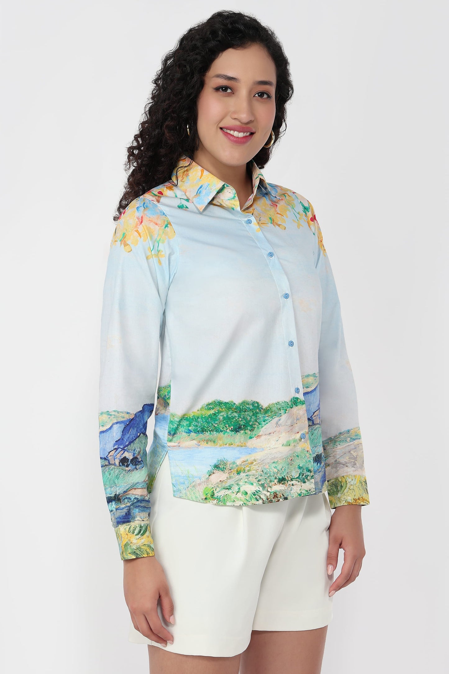 A woman modeling a long-sleeve button-up shirt with a light blue base and a colorful landscape print, paired with white shorts, against a plain white studio background.