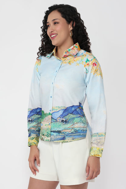 A woman wearing a light blue long-sleeve button-up shirt with a colorful landscape and floral print, paired with white shorts, against a plain light gray background.