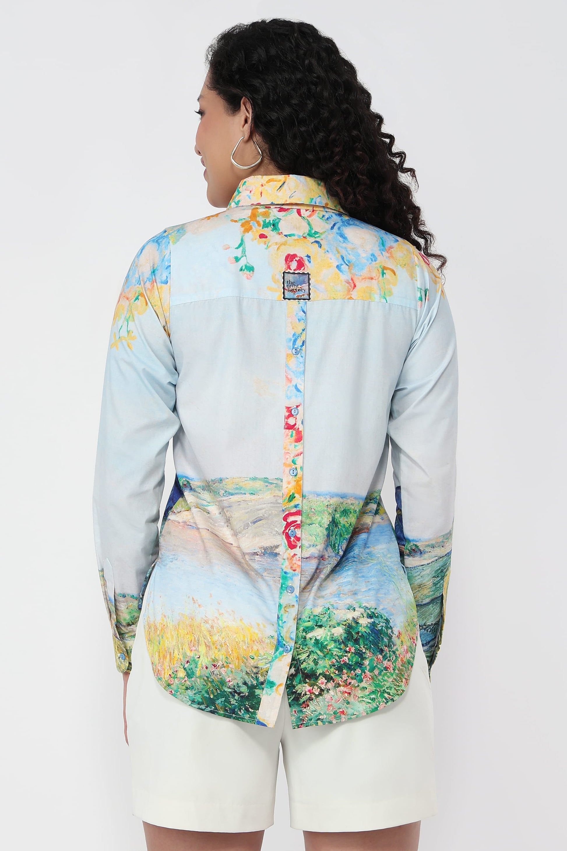A woman shown from the back wearing a long-sleeve button-up shirt with a light blue base and a multicolor landscape print, paired with white shorts against a plain white background.