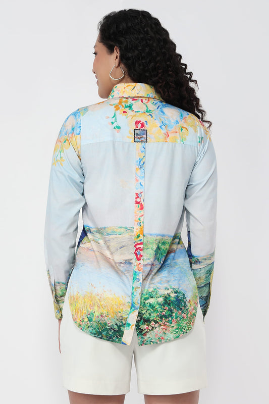 A woman shown from the back wearing a long-sleeve button-up shirt with a light blue base and a multicolor landscape print, paired with white shorts against a plain white background.