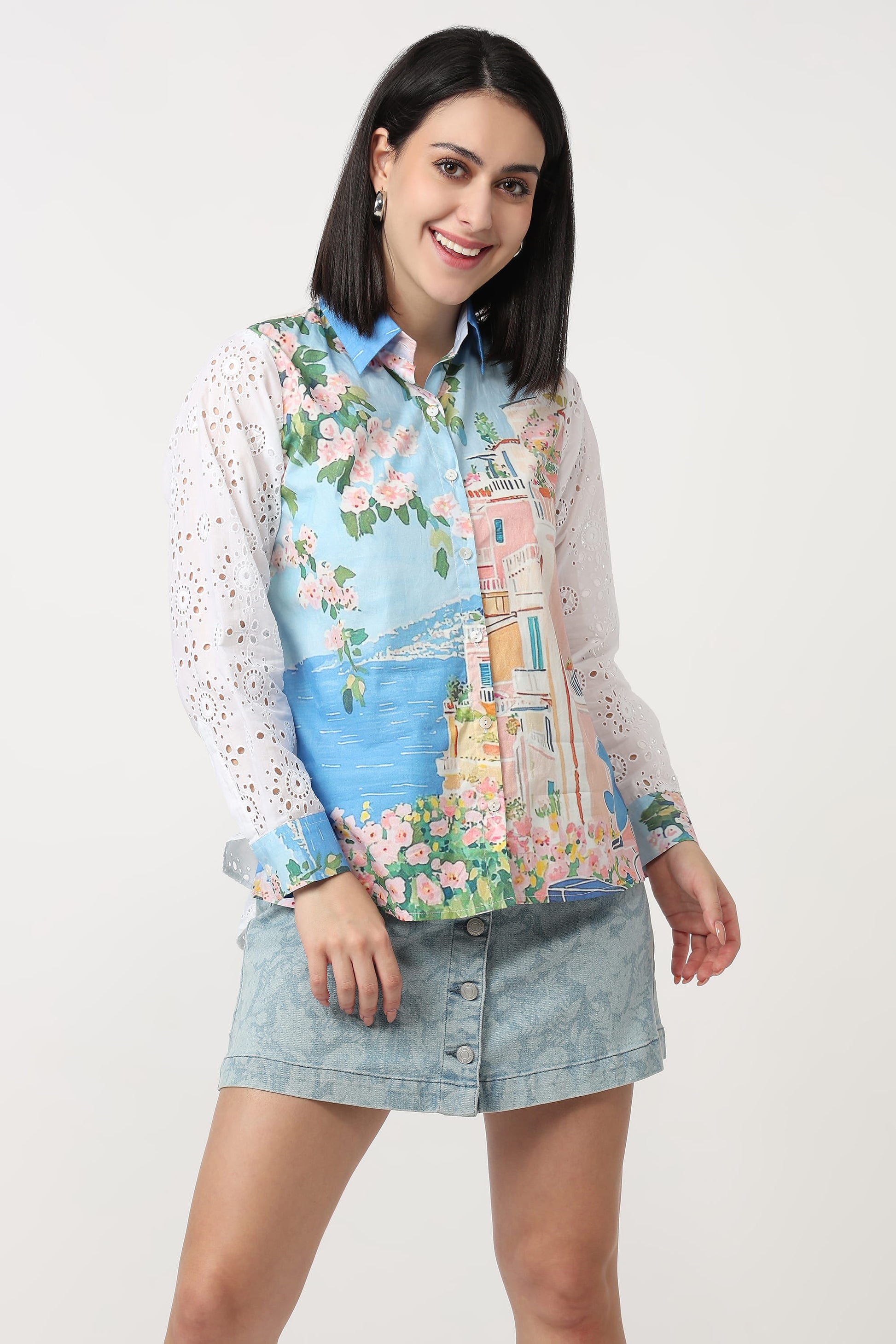 A woman modeling a long-sleeve button-up shirt with a colorful scenic print of a coastal town and a light blue denim mini skirt, set against a plain white studio background.