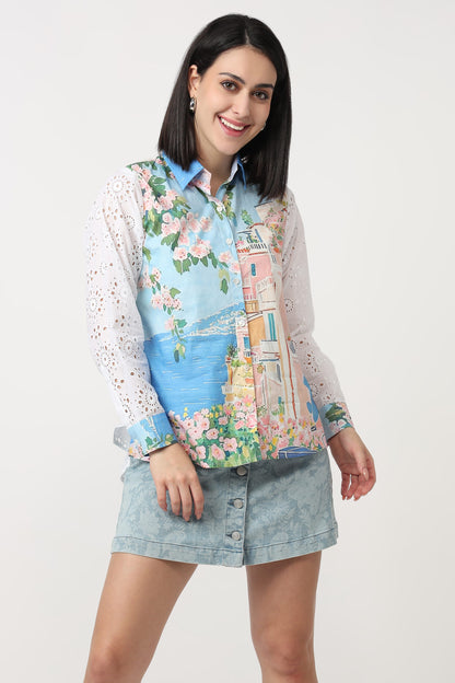 A woman modeling a long-sleeve button-up shirt with a colorful scenic print of a coastal town and a light blue denim mini skirt, set against a plain white studio background.