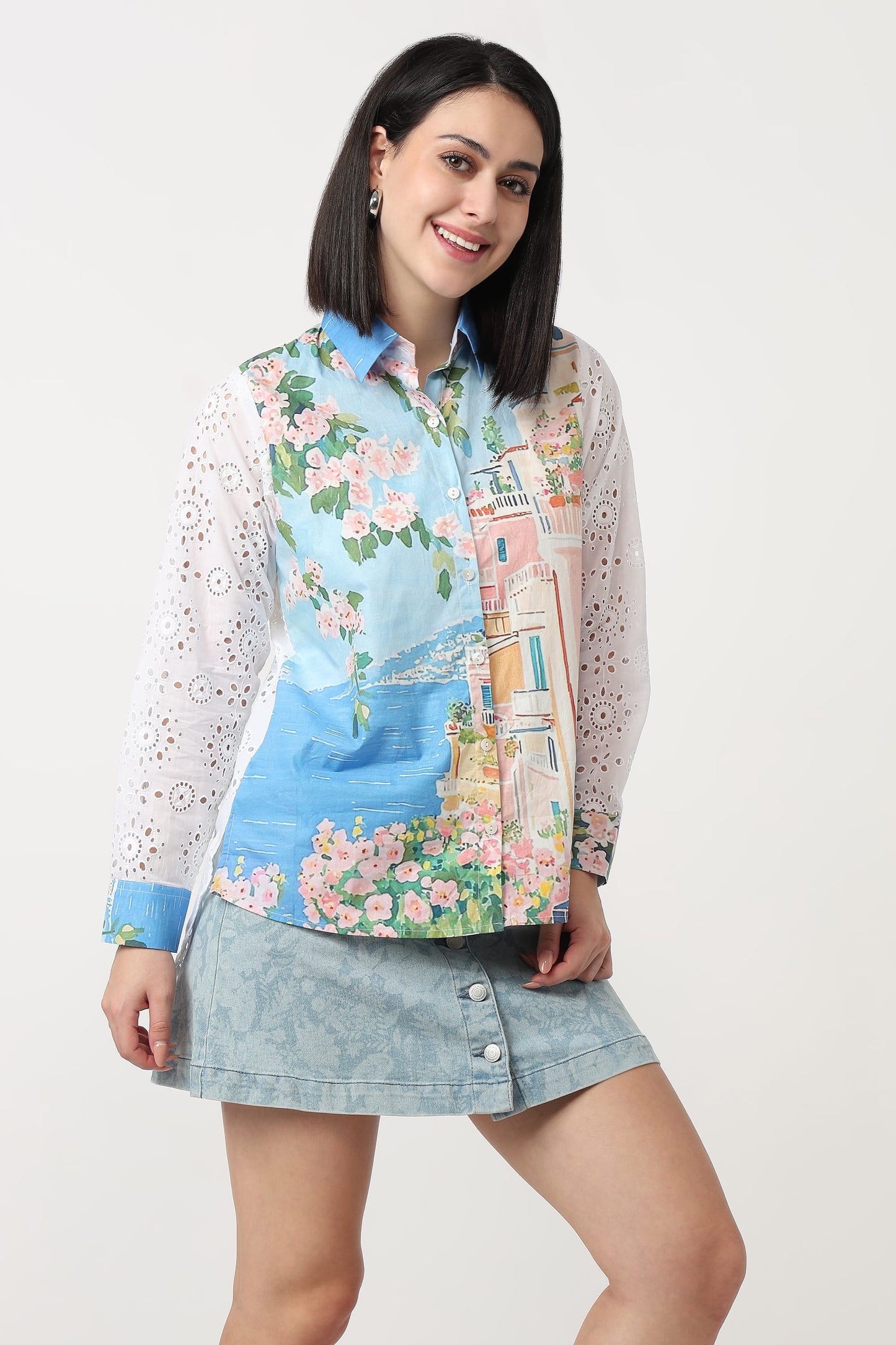A woman modeling a long-sleeve button-up shirt with a colorful scenic print featuring blue water, pink flowers, and a building, paired with a light blue denim mini skirt against a plain light gray background.