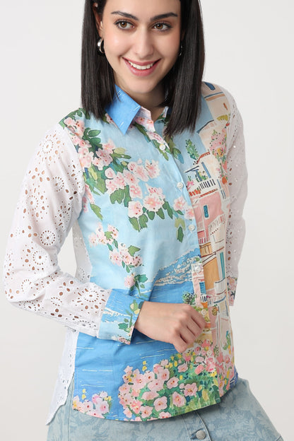 A woman wearing a long-sleeve button-up shirt with a light blue base, featuring a colorful coastal town print with pink flowers, green leaves, and white eyelet lace sleeves.