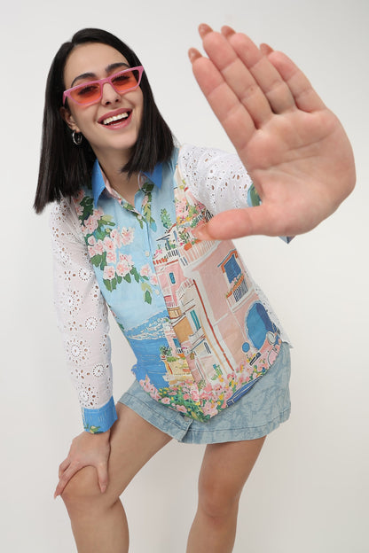 A woman wearing a colorful long-sleeve button-up shirt with a pastel Mediterranean village print and white lace sleeves, paired with denim shorts, posing with one hand raised in front of her face against a plain white background.