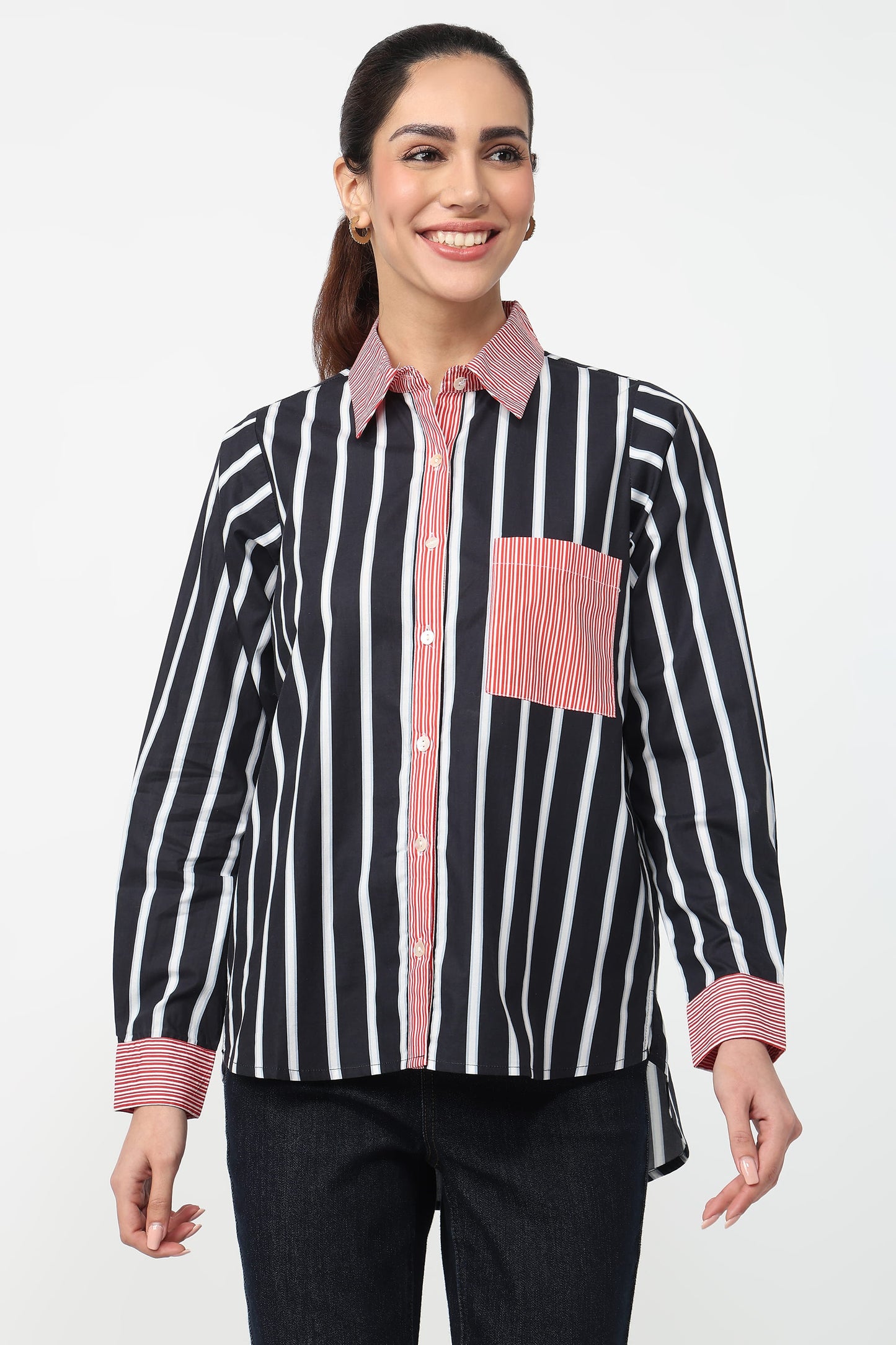 A woman modeling a long-sleeve button-up shirt with vertical black and white stripes, featuring a pink and white striped collar, placket, cuffs, and a matching pink striped chest pocket, paired with dark jeans against a light gray background.