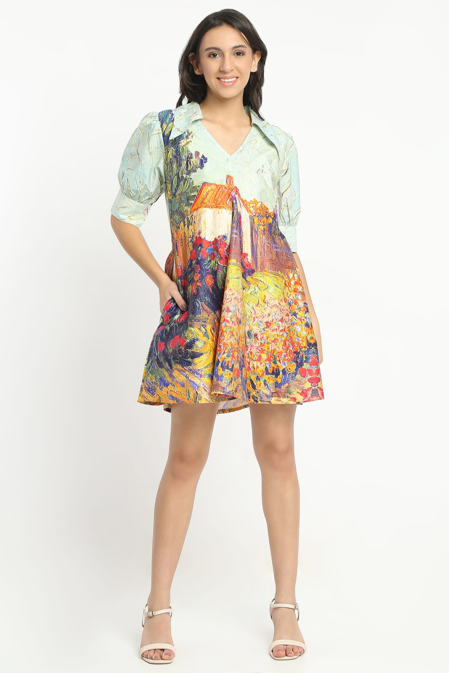 A woman wearing  a short puff-sleeve dress with a multicolour floral and landscape print, paired with beige strappy high-heeled sandals, against a plain white studio background.