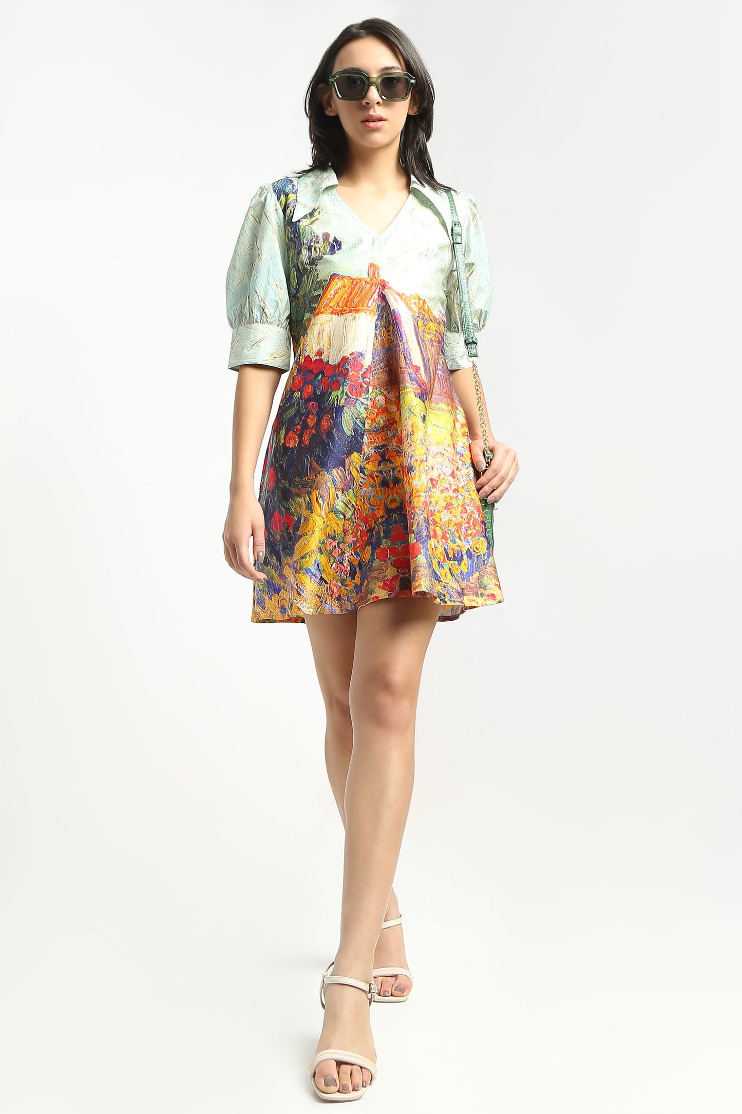 A woman wearing  a short puff-sleeve dress with a multicolour floral and landscape print, paired with beige strappy high-heeled sandals, against a plain white studio background.