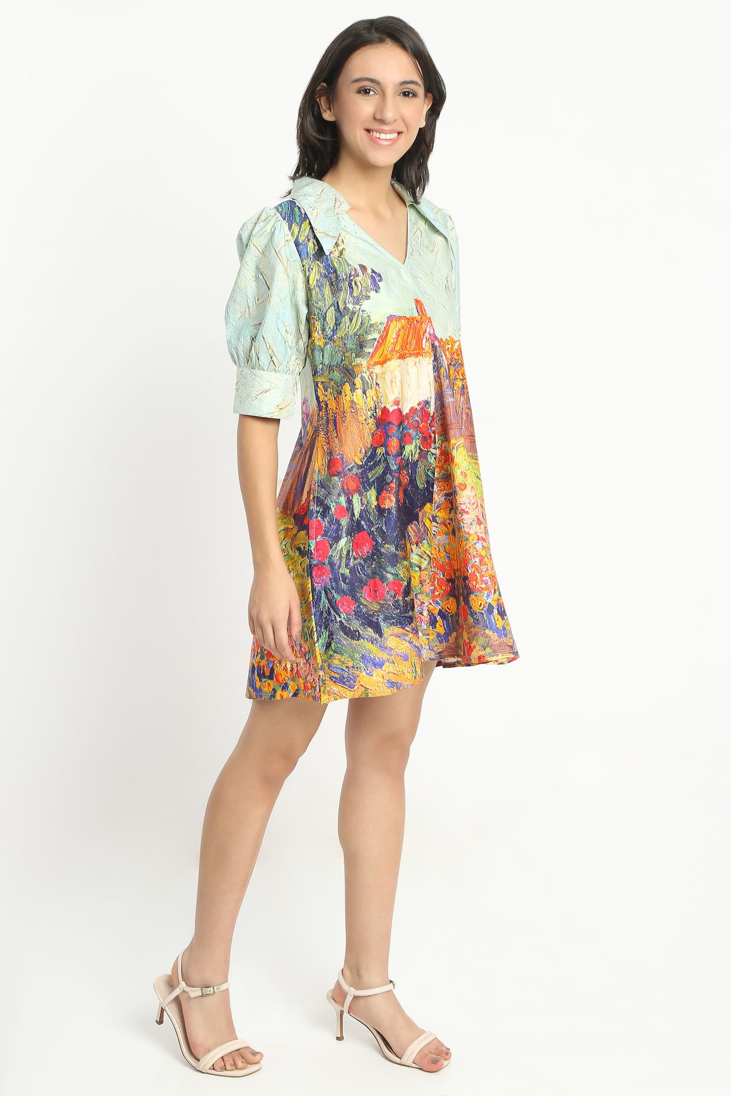 A woman wearing  a short puff-sleeve dress with a multicolour floral and landscape print, paired with beige strappy high-heeled sandals, against a plain white studio background.