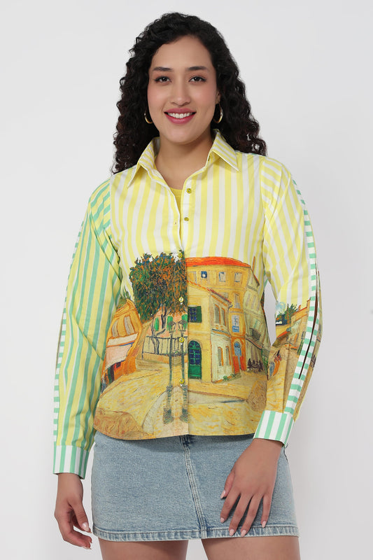 A woman modeling a long-sleeve button-up shirt with yellow and green vertical stripes on the upper body and sleeves, featuring a printed Van Gogh-style village scene on the lower front, paired with a denim skirt against a light gray background.