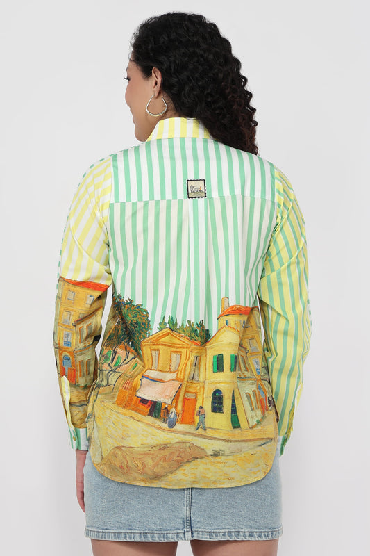A woman shown from the back wearing a long-sleeve button-up shirt with a light green and yellow striped upper back and shoulders, and a Van Gogh-style village painting print across the lower back and sleeves. She is also wearing a denim skirt against a plain light gray studio background.