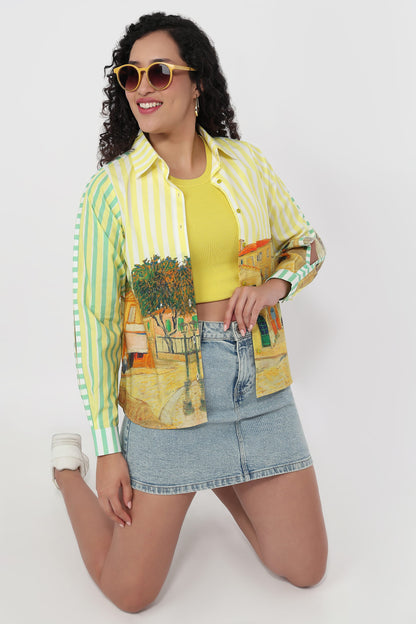 A woman kneels against a light gray studio background wearing a long-sleeve button-up shirt with yellow and mint green vertical stripes on the sleeves and a multicolor artistic print on the front, over a yellow crop top and a light blue denim mini skirt.