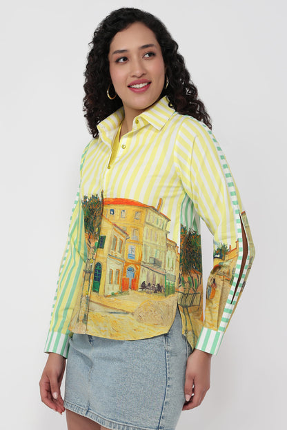 A woman modeling a long-sleeve button-up shirt with yellow and green vertical stripes and a large Van Gogh-style village scene print across the front and sleeves, paired with a denim skirt against a light gray studio background.