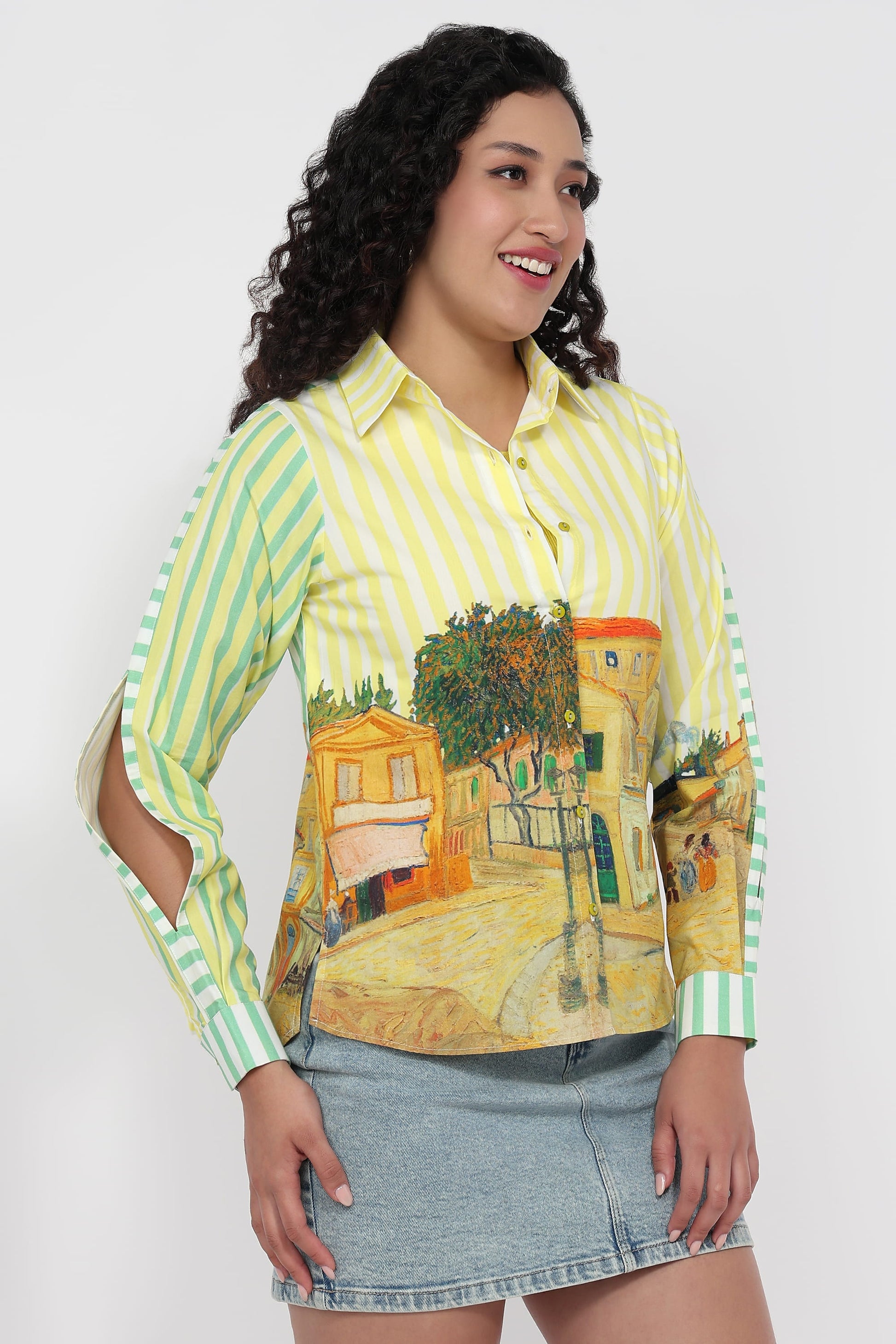A woman modeling a long-sleeve button-up shirt with a yellow and green striped base and a Van Gogh-inspired village print across the front and sleeves, paired with a denim skirt against a light gray background.