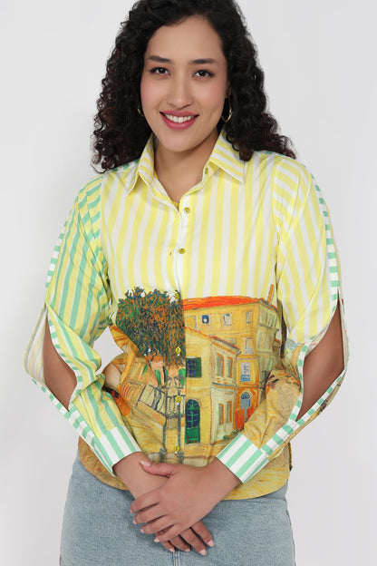 A woman wearing a yellow and green striped button-up shirt with a colorful building and tree graphic across the front, paired with light blue jeans, against a plain light gray background.