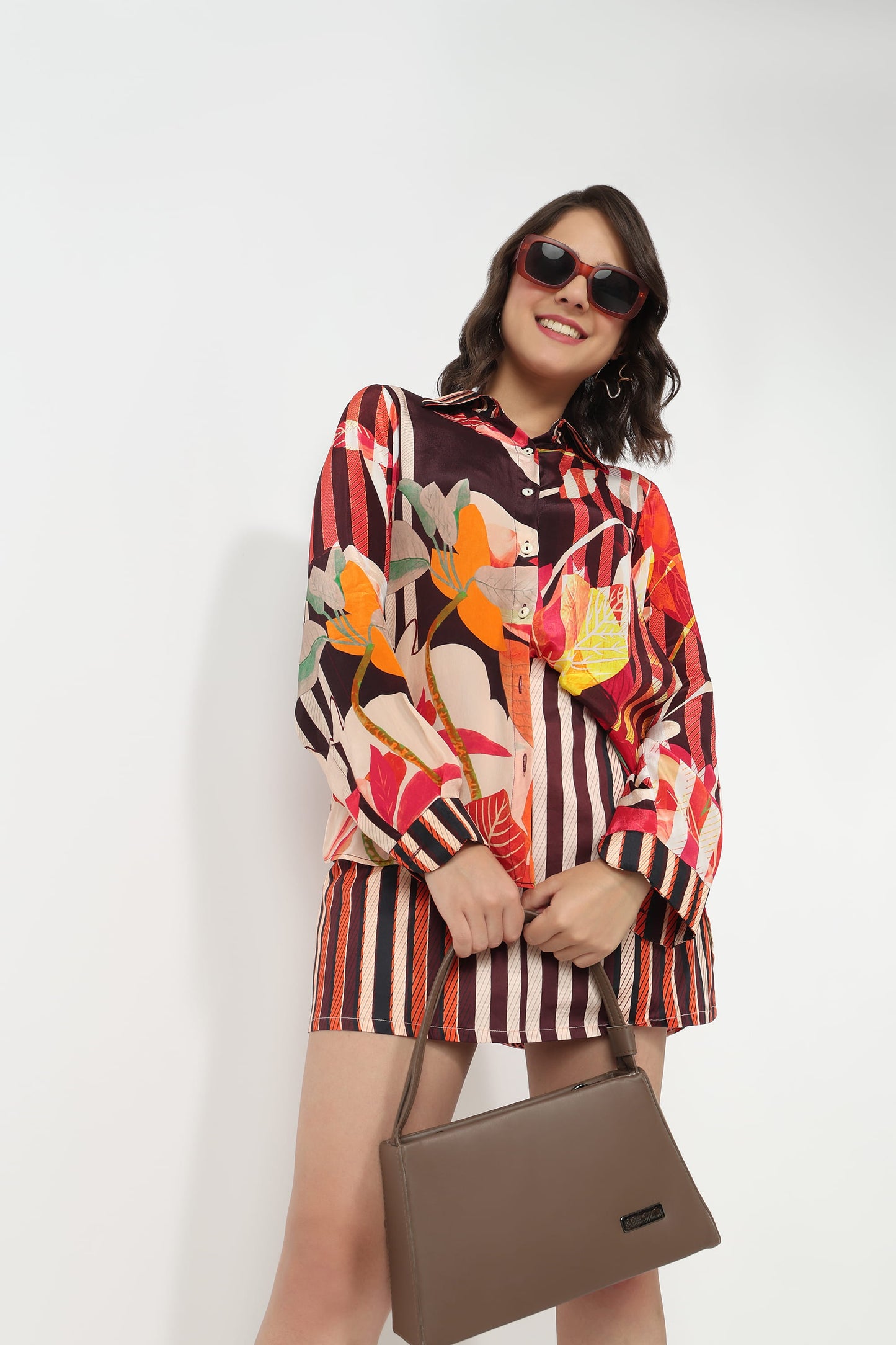 A woman wearing a colorful long-sleeve button-up shirt and matching striped shorts, holding a brown handbag against a plain white background.