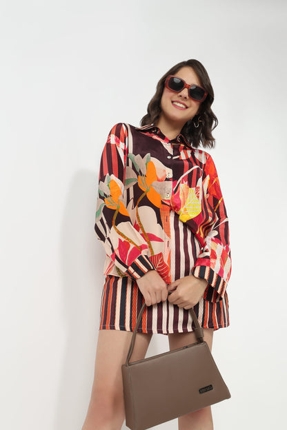 A woman wearing a colorful long-sleeve button-up shirt and matching striped shorts, holding a brown handbag against a plain white background.