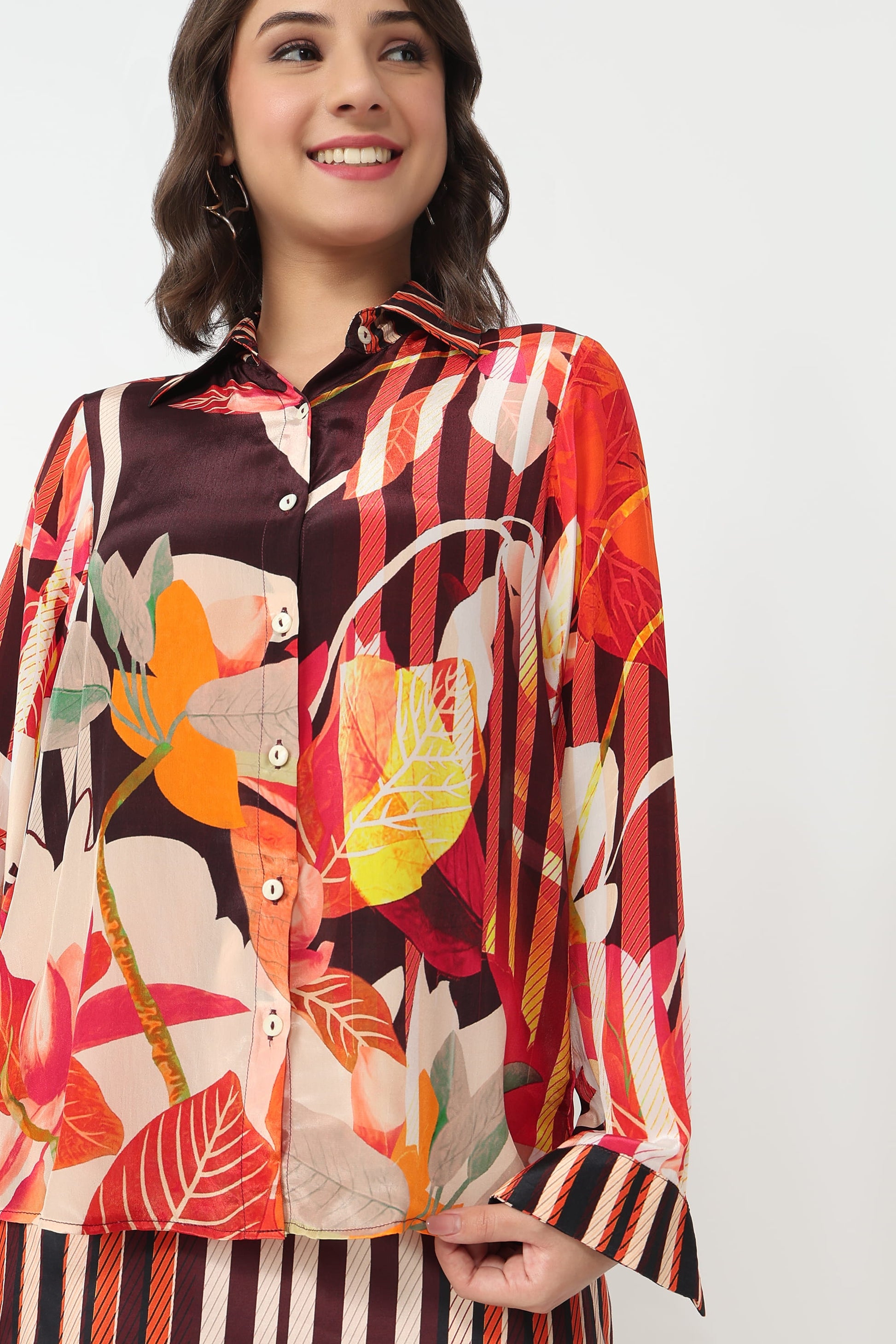 A woman wearing a long-sleeve button-up shirt with a vibrant multicolor botanical and striped pattern, paired with matching striped bottoms, against a plain light gray background.