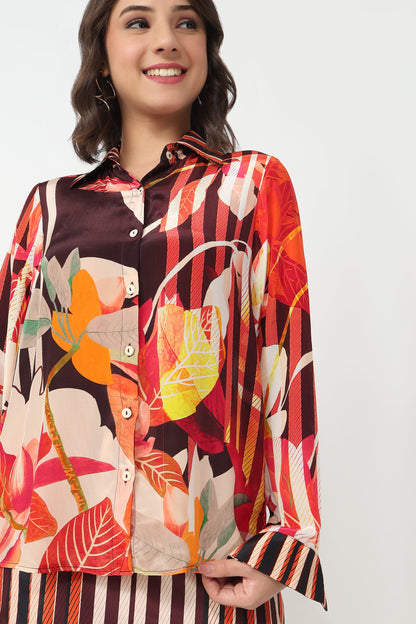 A woman wearing a long-sleeve button-up shirt with a vibrant multicolor botanical and striped pattern, paired with matching striped bottoms, against a plain light gray background.