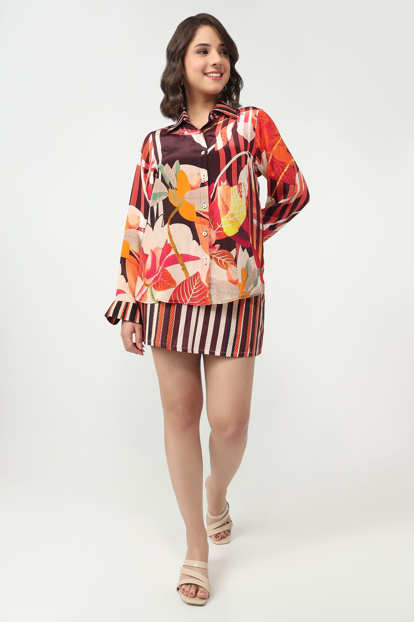 A woman modeling a matching two-piece set featuring a long-sleeve collared shirt with a colorful tropical floral print and striped cuffs, paired with a matching striped mini skirt.