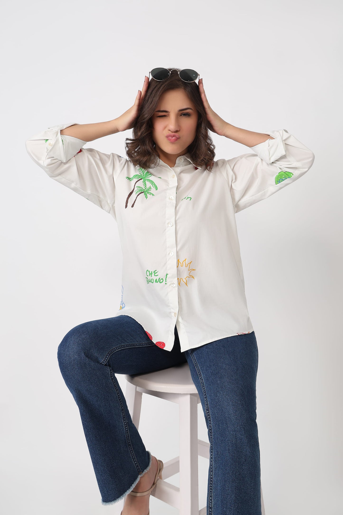 Woman wearing a white shirt with green palm tree designs and blue jeans, sitting on a white stool against a white background.