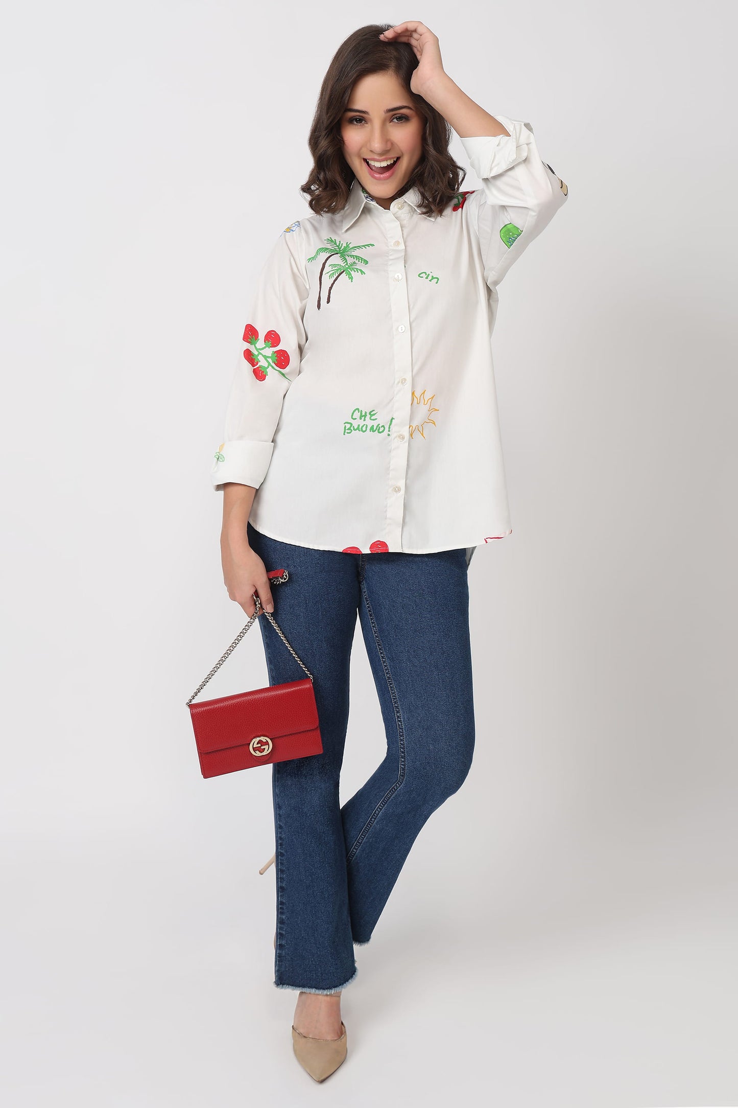 Woman wearing a white shirt with colorful embroidery and blue jeans, holding a red handbag on a white background