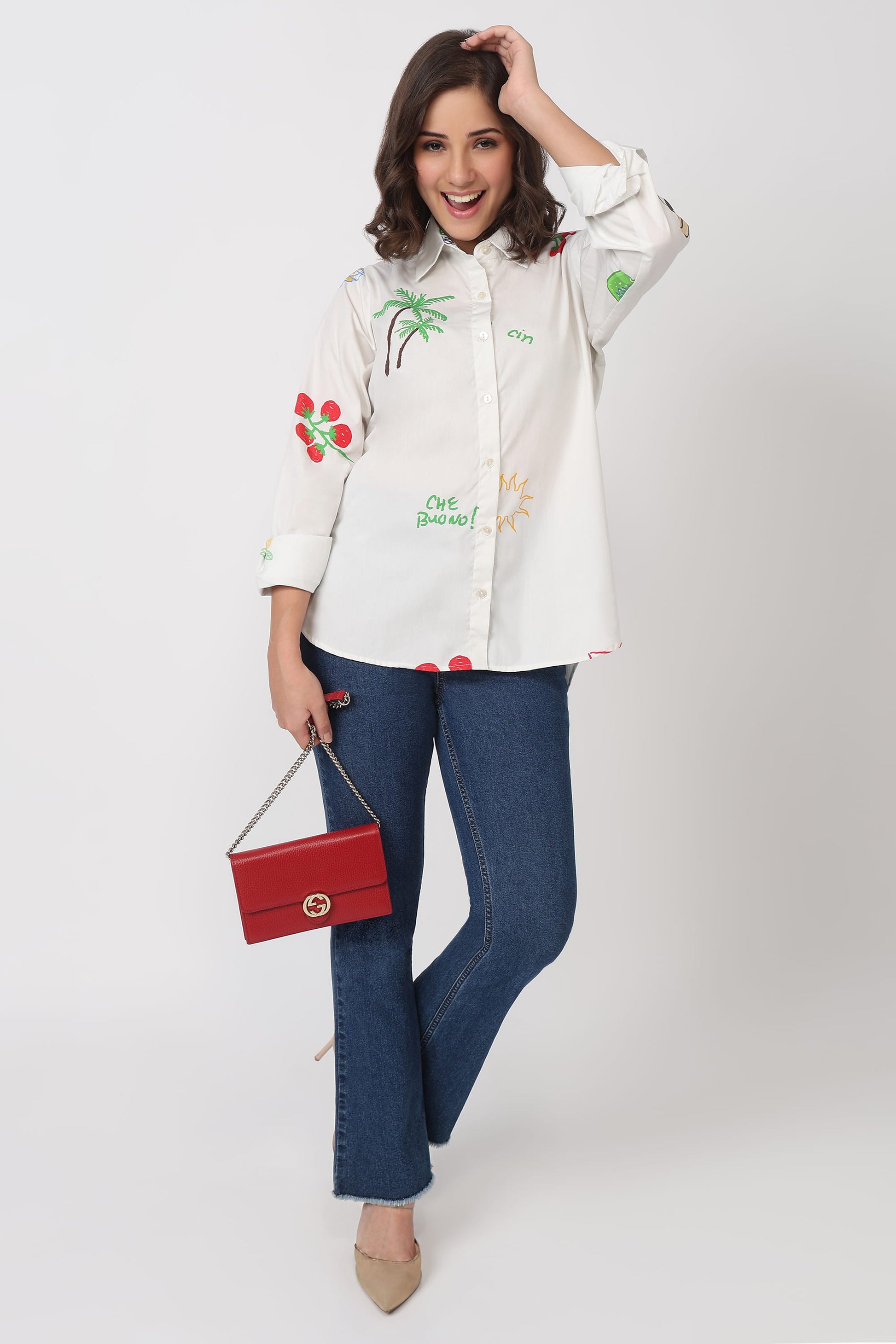 Woman wearing a white shirt with colorful embroidery and blue jeans, holding a red handbag on a white background