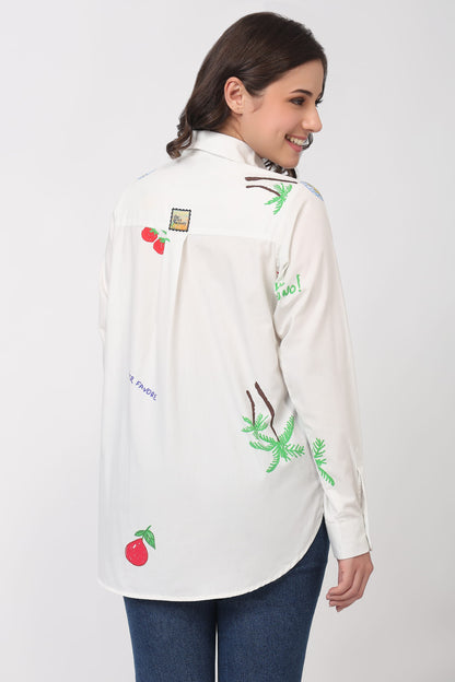A woman wearing a white long-sleeve button-up shirt with colorful tropical and fruit print, paired with blue jeans, shown from a three-quarter back view against a light gray studio background.