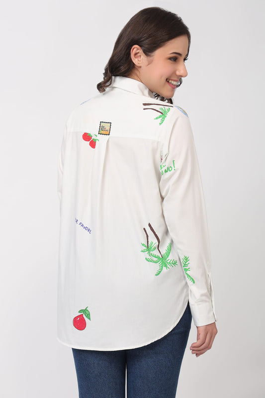 A woman wearing a white long-sleeve button-up shirt with colorful tropical and fruit print, paired with blue jeans, shown from a three-quarter back view against a light gray studio background.