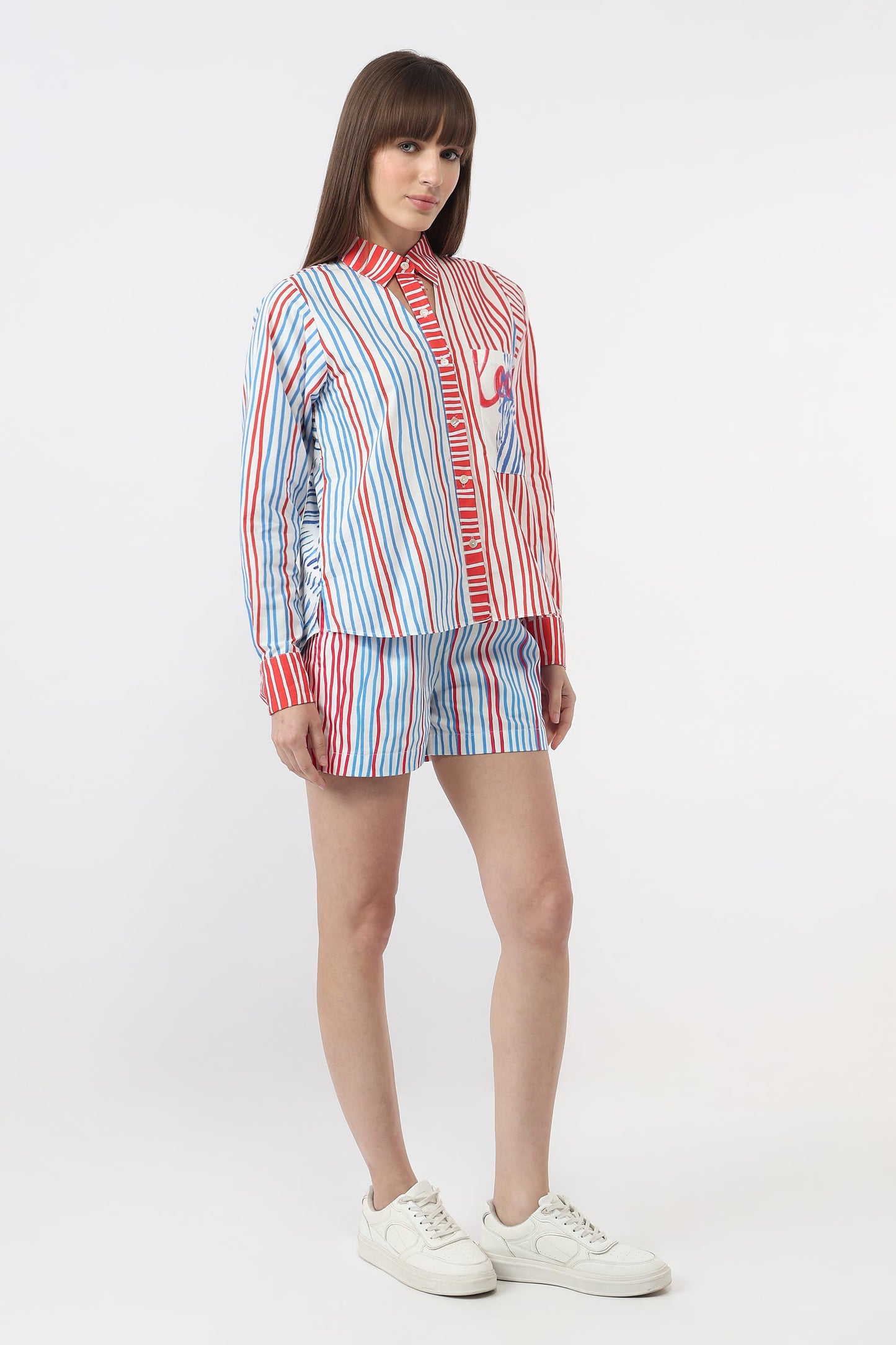 A model wearing a matching striped shirt and shorts set in red, white, and blue, paired with white sneakers, against a plain light gray studio background.