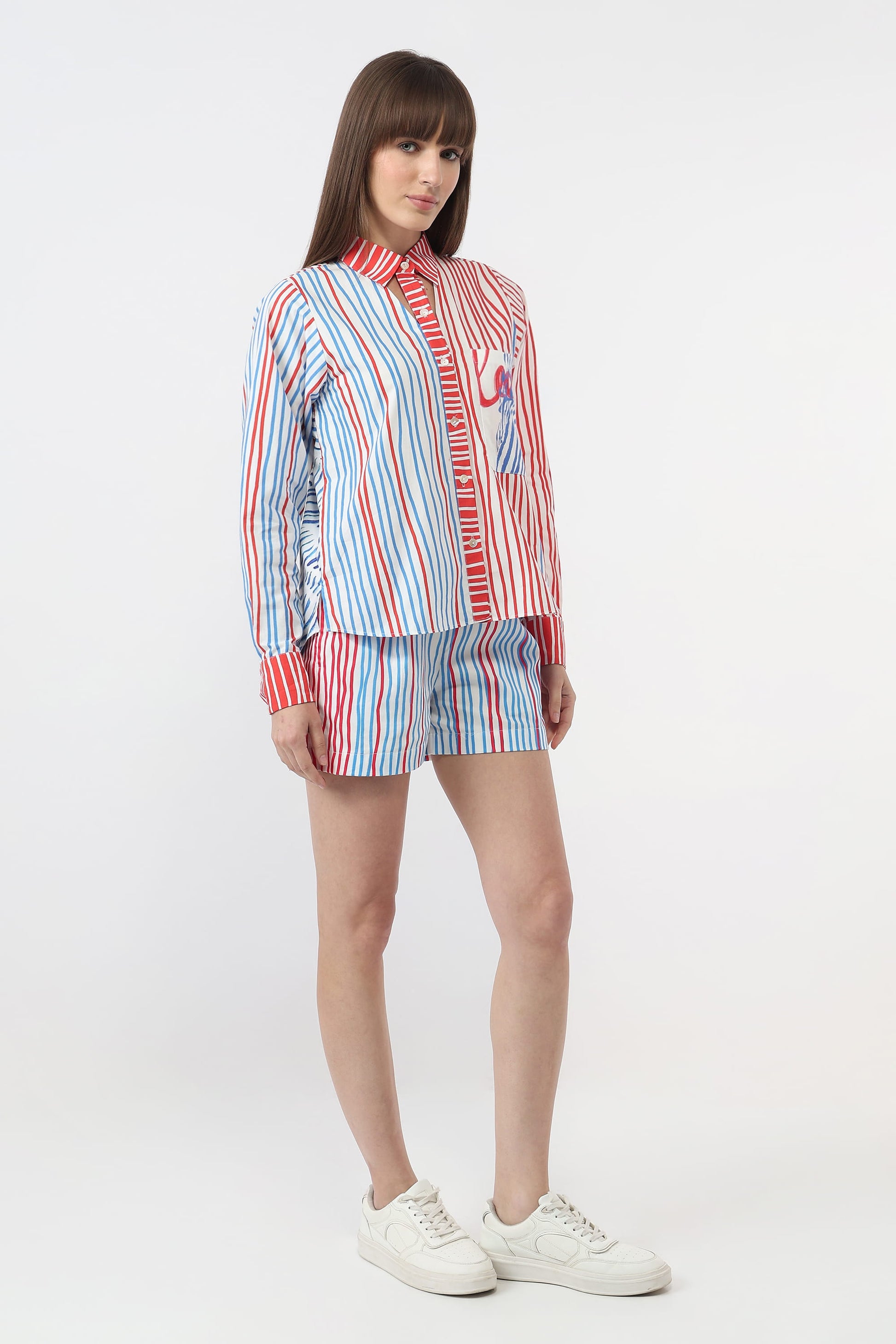 A model wearing a matching striped shirt and shorts set in red, white, and blue, paired with white sneakers, against a plain light gray studio background.