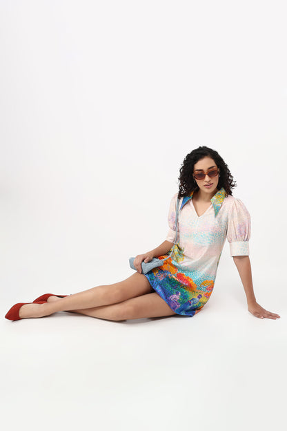 A woman wearing a short, colorful printed dress with puff sleeves and sunglasses sits on a white studio floor, holding a gray clutch.