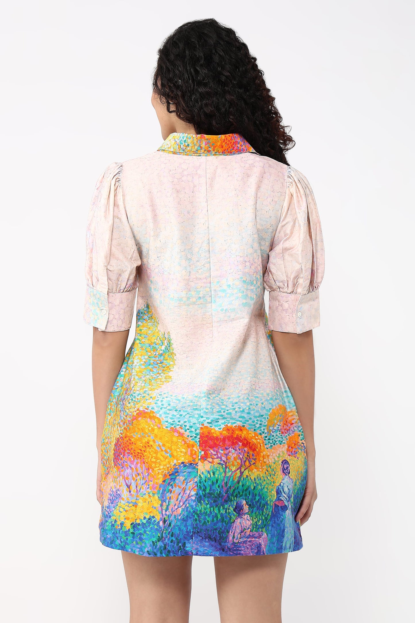 A woman viewed from the back wearing a short puff-sleeve dress with a pastel pink base and a colorful landscape print in blue, yellow, orange, and purple at the bottom.