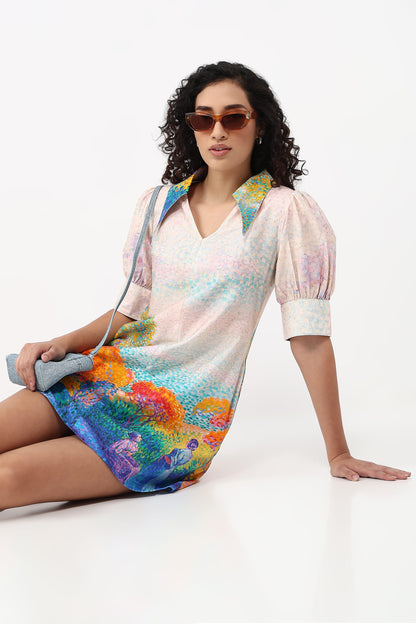 A woman wearing a short puff-sleeve mini dress with a multicolor abstract landscape print, seated against a plain white studio background.