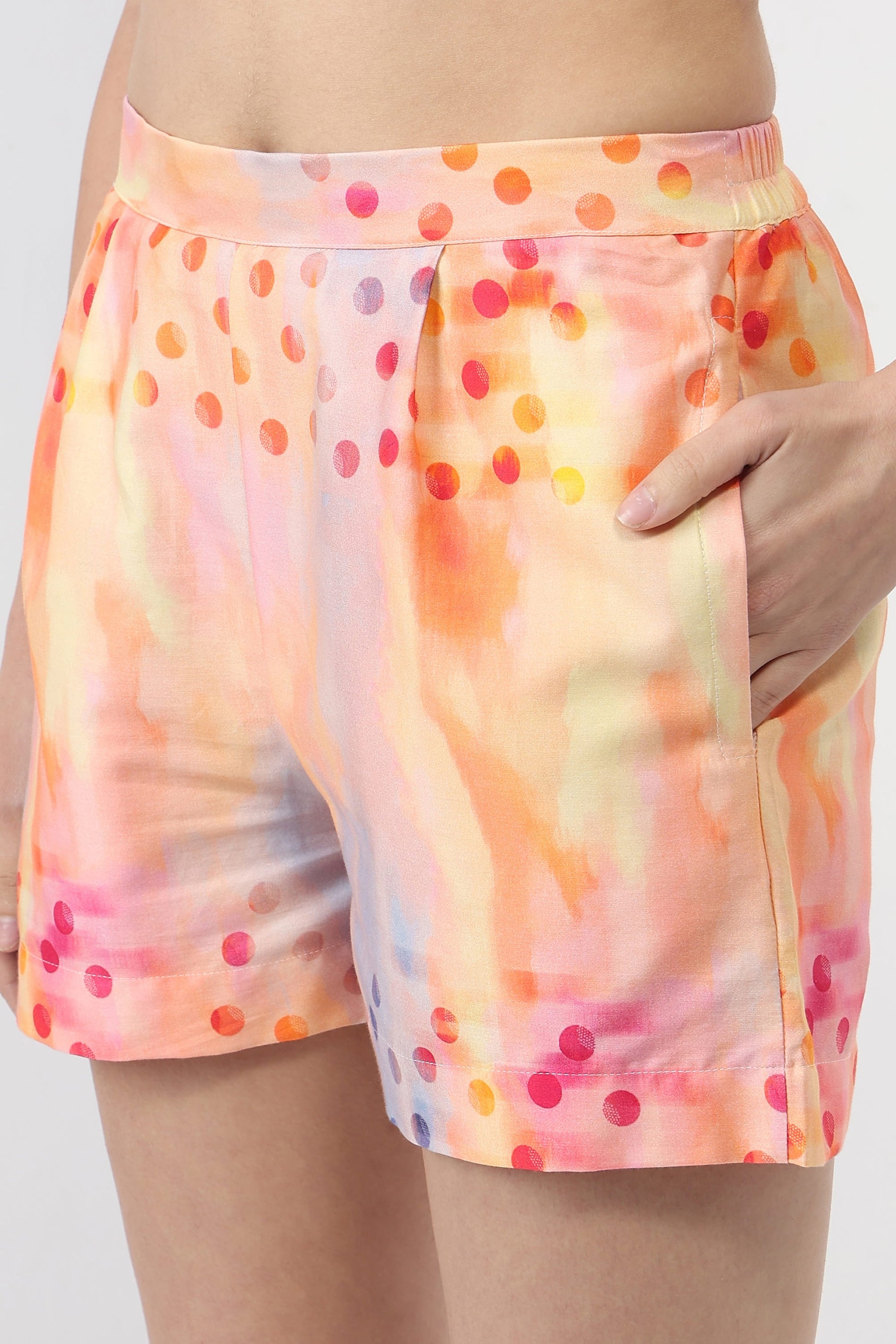 A close-up of a model wearing high-waisted shorts with a pastel watercolor wash and scattered multicolor polka dots in orange, pink, yellow, and purple.