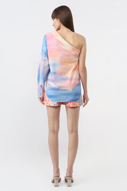 A woman shown from the back wearing a one-shoulder short cover-up with a pastel watercolor print in shades of blue, pink, and peach, paired with matching patterned shorts and heeled sandals, against a plain white studio background.