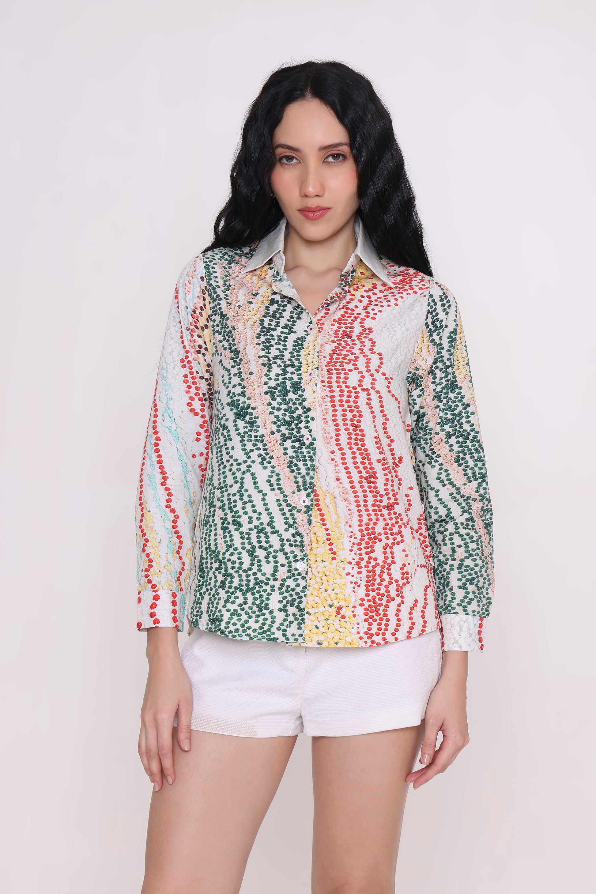 A woman modeling a long-sleeve button-up shirt with a colorful vertical dotted stripe pattern in green, red, yellow, and white, paired with white shorts against a plain light background.